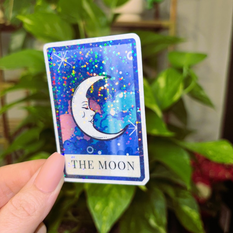 May include: A tarot card sticker featuring a crescent moon with a face, set against a blue, glittery background. The card is labeled "THE MOON" in white text, with a white border.
