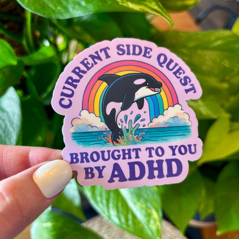 May include: A sticker with a pink background featuring a black and white orca leaping through a rainbow. The text reads "CURRENT SIDE QUEST" and "BROUGHT TO YOU BY ADHD."