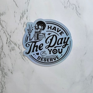 May include: A blue and silver iridescent sticker with a skeleton holding up a peace sign and the text "Have the Day You Deserve."