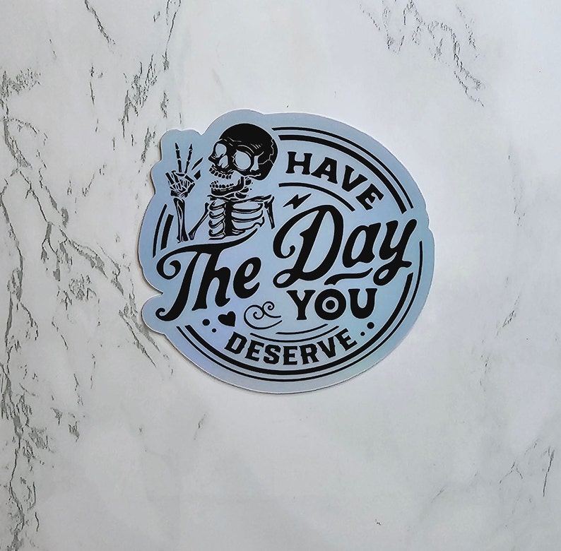 May include: A blue and silver iridescent sticker with a skeleton holding up a peace sign and the text "Have the Day You Deserve."