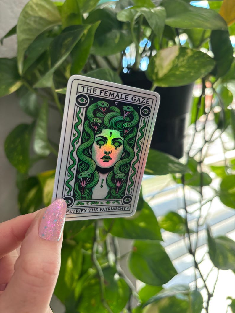 May include: A holographic sticker with a green and white illustration of a woman with snakes for hair. The text "THE FEMALE GAZE" and "PETRIFY THE PATRIARCHY" is printed on the sticker.