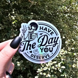May include: A white iridescent sticker with a black skeleton holding up a peace sign and the text "Have the day you deserve".