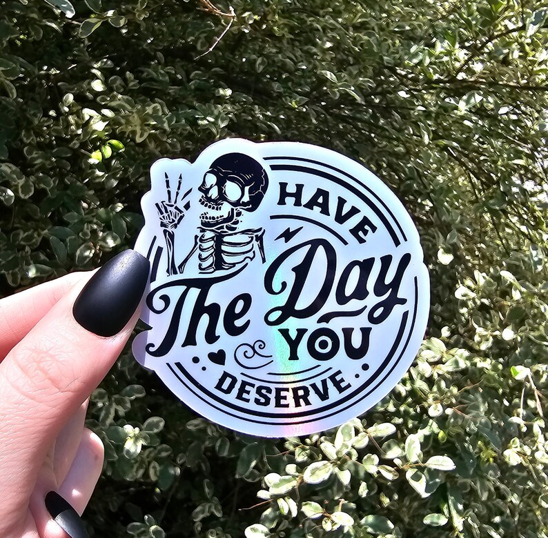 May include: A white iridescent sticker with a black skeleton holding up a peace sign and the text "Have the day you deserve".