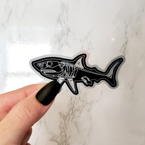 K&ouml;nnte beinhalten: Ein schwarz-wei&szlig;er Aufkleber mit einem Haifisch-Skelett. Der Aufkleber befindet sich auf einem wei&szlig;en Marmoruntergrund.