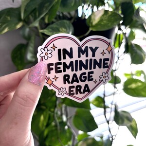 May include: A pink heart-shaped sticker with the text "In My Feminine Rage Era" in black lettering. The sticker has white flowers and gold stars around the edges.