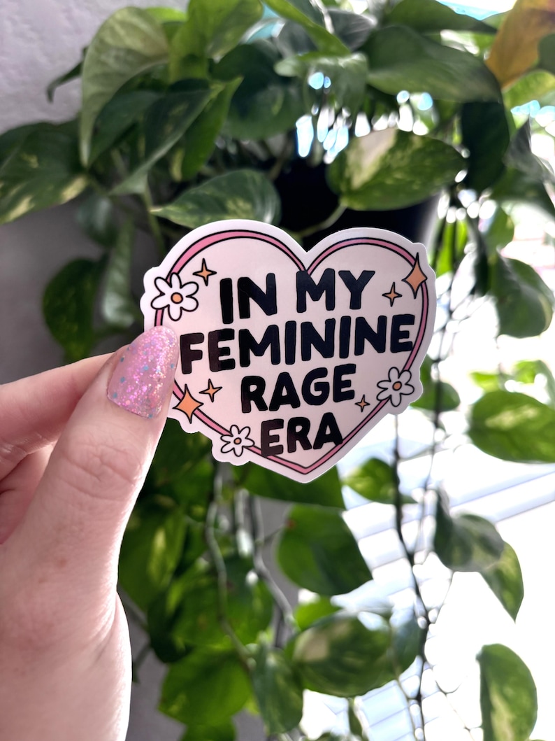 May include: A pink heart-shaped sticker with the text "In My Feminine Rage Era" in black lettering. The sticker has white flowers and gold stars around the edges.