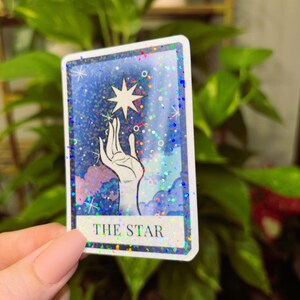 May include: A holographic tarot card with a blue background and a hand reaching up to a star. The card reads "THE STAR" at the bottom.