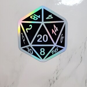 May include: A black and holographic sticker of a twenty-sided die, with white numbers. The die is a dodecahedron shape, with each face displaying a number from 2 to 20. The holographic effect creates a rainbow sheen.