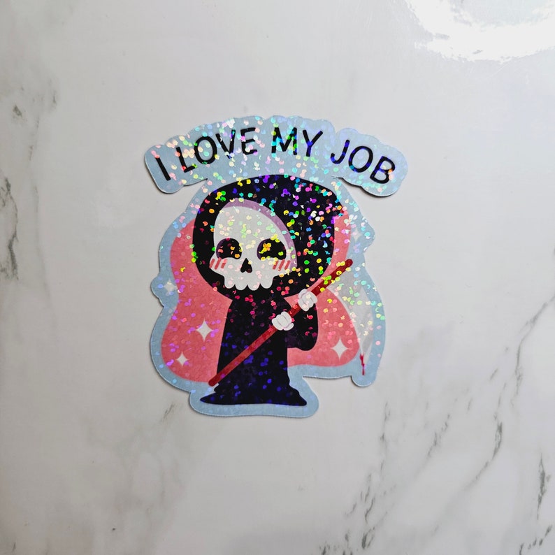 Op de afbeelding: Een holografische sticker met een cartoon Grim Reaper met een zeis, die een zwarte robe draagt en een schedelgezicht heeft. De sticker heeft een roze achtergrond met glitter en de tekst "IK HOUD VAN MIJN WERK".