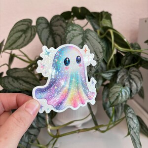 May include: A holographic sticker featuring a cute ghost with a colorful, galaxy-like design. The ghost has big blue eyes and is surrounded by sparkling stars. The sticker is held in front of a green plant.