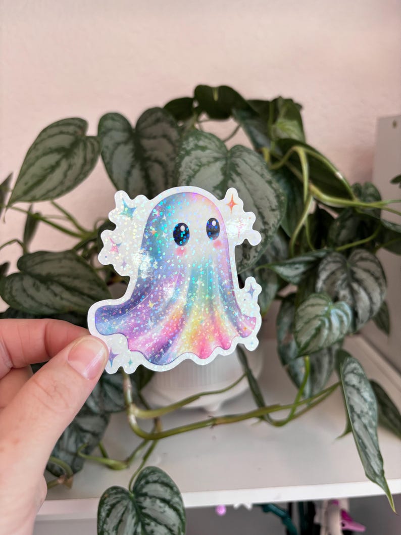 May include: A holographic sticker featuring a cute ghost with a colorful, galaxy-like design. The ghost has big blue eyes and is surrounded by sparkling stars. The sticker is held in front of a green plant.