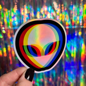 May include: A round sticker featuring an alien face with a black outline. The alien's face is layered with red, yellow, and blue, creating a 3D effect. The sticker has a holographic, iridescent finish.