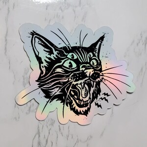 May include: A black and white illustration of a cat with five eyes and an open mouth, printed on a holographic sticker. The cat's eyes are outlined in white, and the sticker has a rainbow iridescent finish.