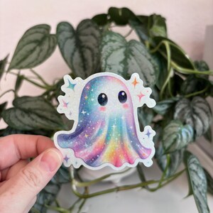 May include: A colorful sticker featuring a cartoon ghost with a rainbow design. The ghost has a sparkly, iridescent finish and is surrounded by star-shaped accents. The sticker is held in front of a background of green plants.