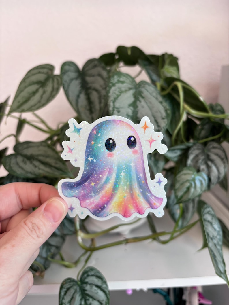 May include: A colorful sticker featuring a cartoon ghost with a rainbow design. The ghost has a sparkly, iridescent finish and is surrounded by star-shaped accents. The sticker is held in front of a background of green plants.