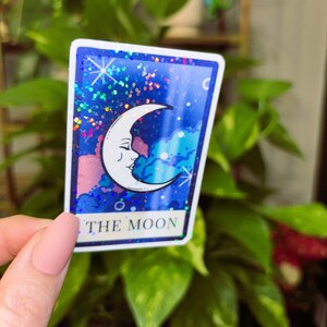 May include: A rectangular sticker featuring a crescent moon with a face, set against a starry blue background with pink and blue clouds. The text "THE MOON" is at the bottom. The sticker has a glittery border.