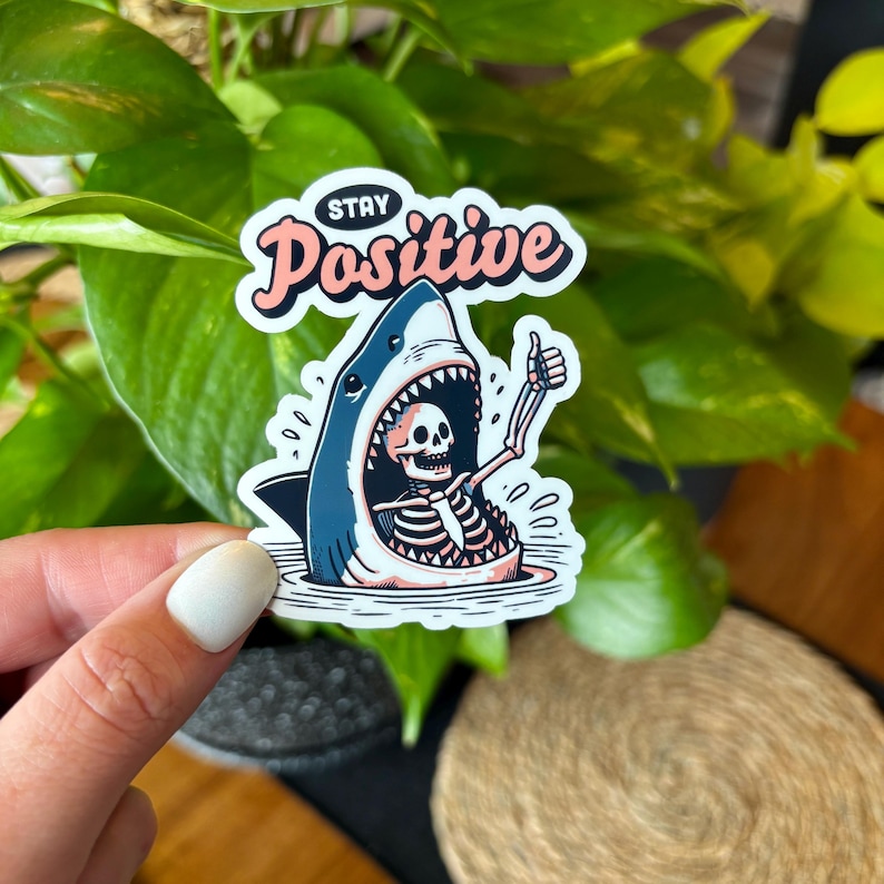 May include: A sticker featuring a cartoon shark with a skeleton inside, giving a thumbs-up. The shark is blue and white, with the skeleton in black and white. The sticker has the words "Stay Positive" in pink and white.