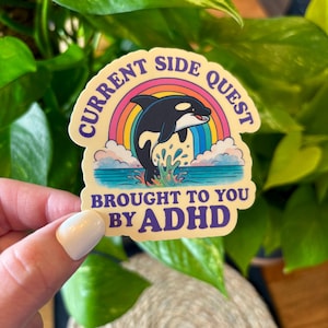 May include: A sticker with a cartoon orca leaping from the ocean, set against a rainbow and clouds. The text reads "CURRENT SIDE QUEST BROUGHT TO YOU BY ADHD" in a playful font. The sticker has a cream-colored background.
