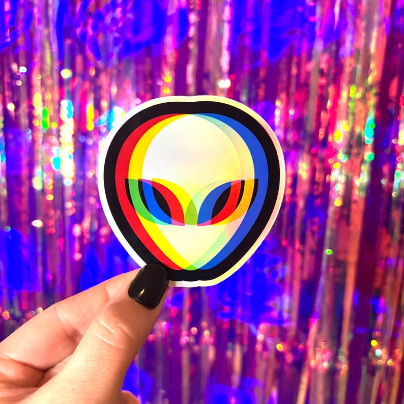May include: A circular sticker featuring an alien face with a white background, outlined in black. The alien's eyes are red, yellow, and blue, creating a 3D effect. The sticker is held against a shimmering, purple background.