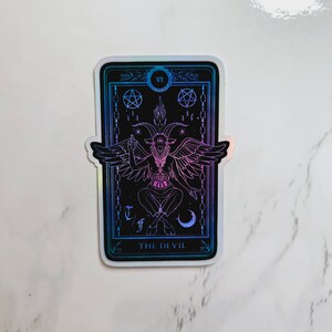 May include: A holographic sticker featuring a tarot card with the Devil, a horned figure with wings, and the text "THE DEVIL" at the bottom.