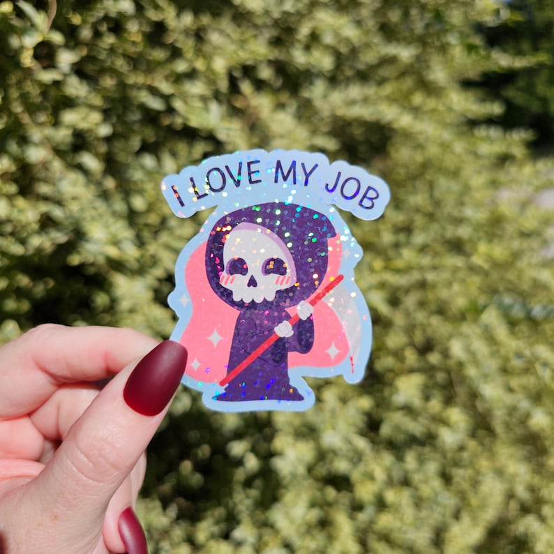 Op de afbeelding: Een holografische sticker met een cartoon Grim Reaper met een roze halo en de tekst "Ik hou van mijn werk".