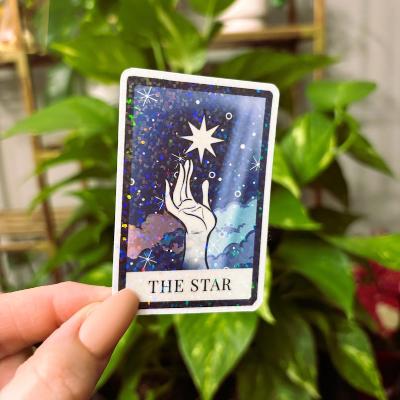 May include: A holographic sticker featuring a hand reaching towards a star with a blue, purple, and white background. The sticker has the text "THE STAR" at the bottom.