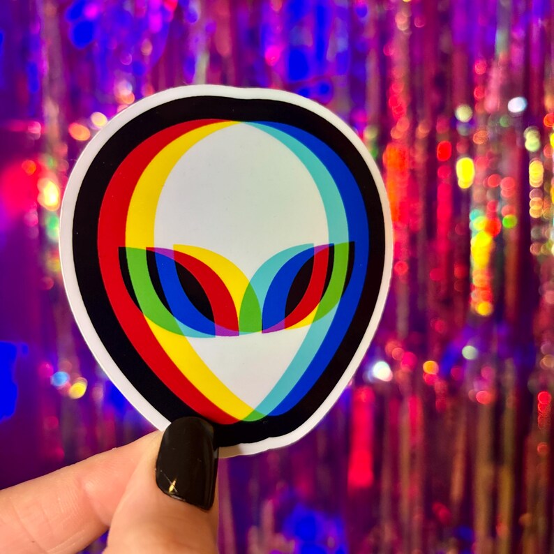 May include: A white alien sticker with a black border and a colorful 3D effect. The alien's eyes are red, yellow, and blue. The background is a blurred, iridescent, multi-colored surface.