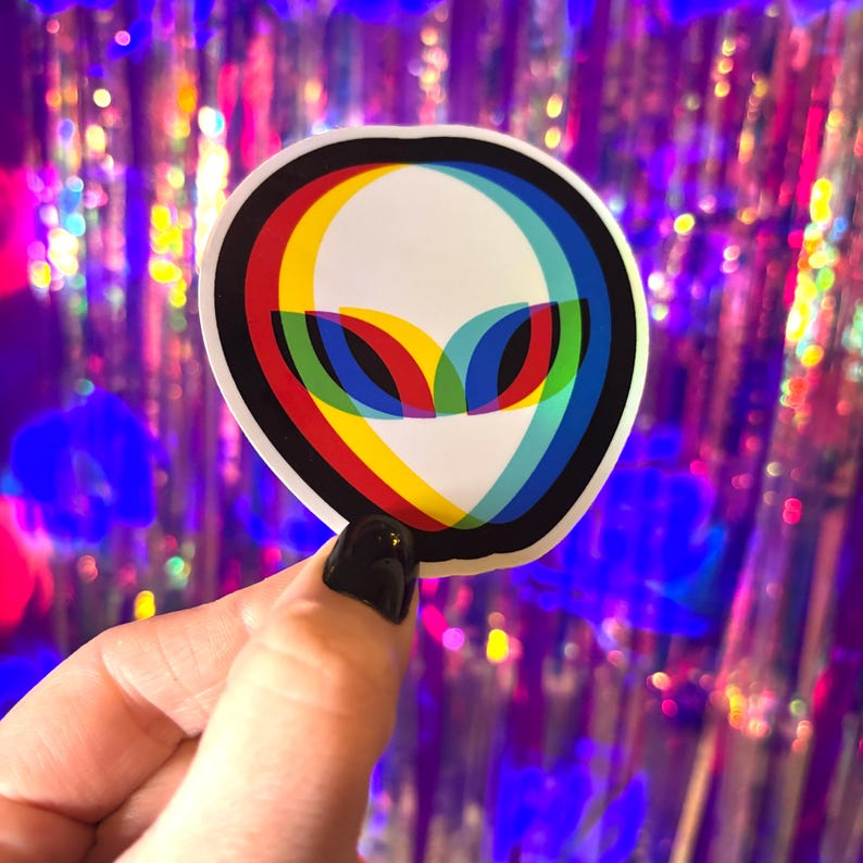 May include: A white alien head sticker with a black border and a 3D effect. The alien's eyes are red, yellow, and blue. The sticker is held by a hand with a black painted nail.