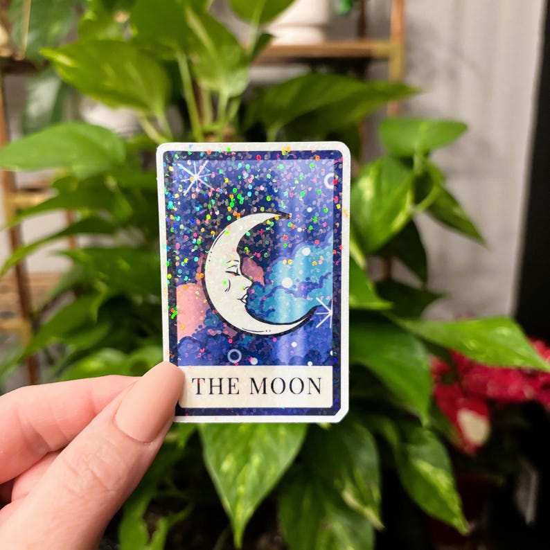 May include: A rectangular sticker featuring a crescent moon with a face, set against a starry, glittery background. The card has a blue border and the words "THE MOON" at the bottom. The sticker is held in front of a green leafy background.