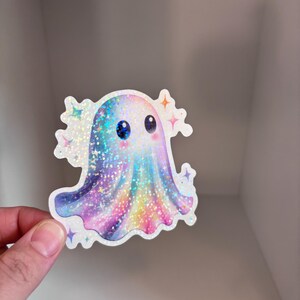May include: A holographic sticker featuring a cute ghost with big blue eyes and a colorful, rainbow-like design. The ghost has a sparkly, glittery appearance and is surrounded by star-shaped accents. The sticker is held by a hand.