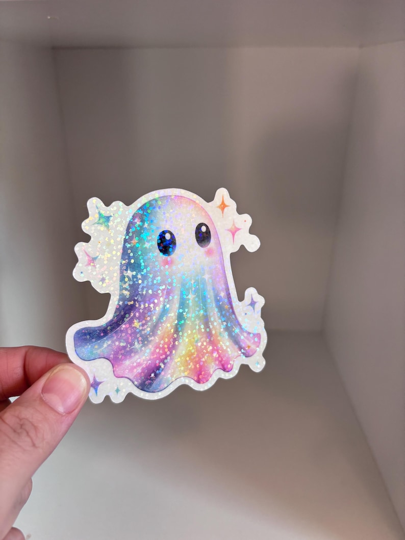 May include: A holographic sticker featuring a cute ghost with big blue eyes and a colorful, rainbow-like design. The ghost has a sparkly, glittery appearance and is surrounded by star-shaped accents. The sticker is held by a hand.