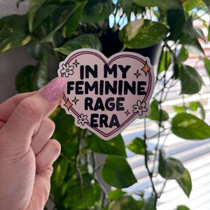 May include: A pink heart-shaped sticker with a white outline and black text that reads "In My Feminine Rage Era". The sticker has small white flowers and gold stars around the text.