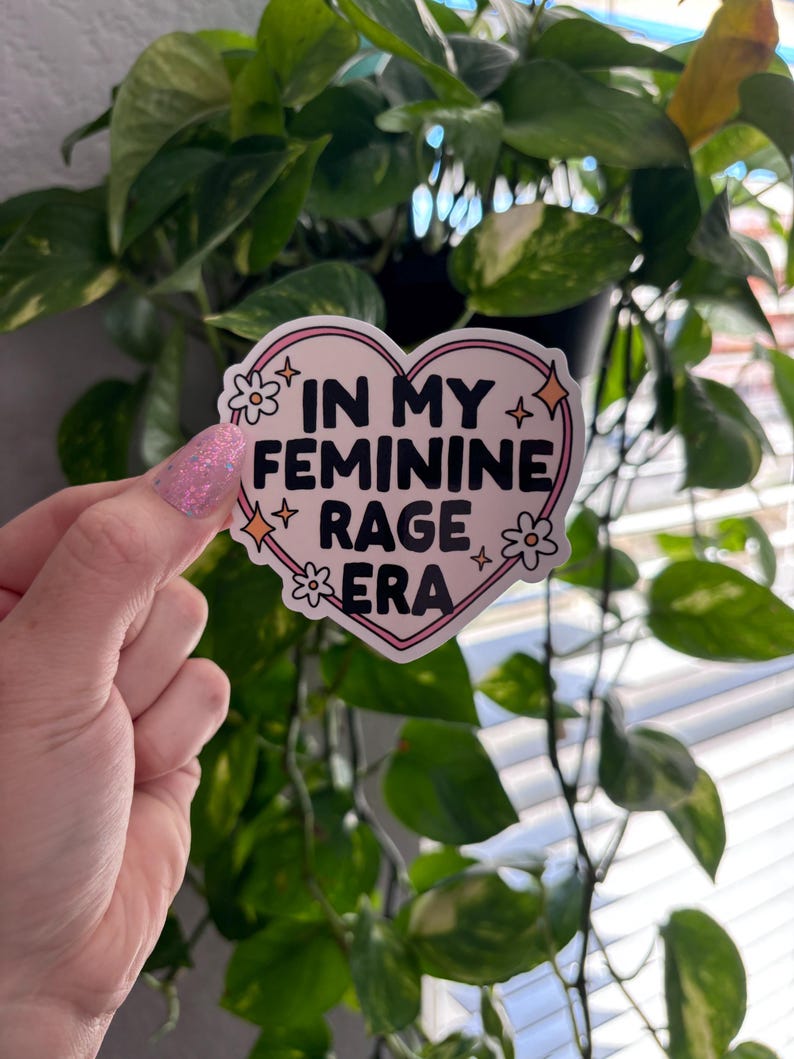 May include: A pink heart-shaped sticker with a white outline and black text that reads "In My Feminine Rage Era". The sticker has small white flowers and gold stars around the text.