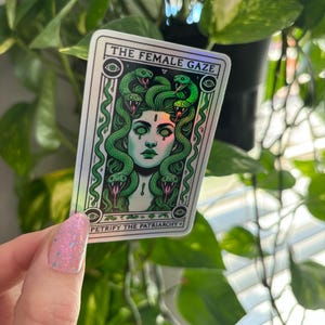 May include: A holographic sticker with a green and white design. The sticker features a woman with snakes for hair and the text "THE FEMALE GAZE" and "PETRIFY THE PATRIARCHY".