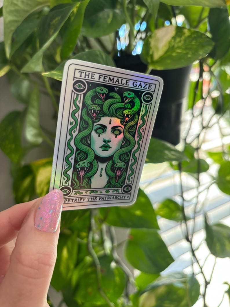 May include: A holographic sticker with a green and white design. The sticker features a woman with snakes for hair and the text "THE FEMALE GAZE" and "PETRIFY THE PATRIARCHY".