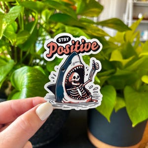 May include: A sticker featuring a cartoon shark with a skeleton inside, giving a thumbs-up. The sticker has the words "Stay Positive" in a cursive font. The shark is blue and white, and the skeleton is white.