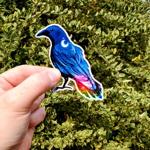 May include: A blue crow sticker with a crescent moon and stars on its chest. The crow's tail feathers are iridescent with a rainbow of colors.