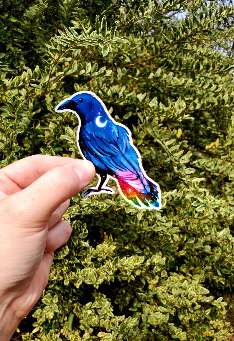 May include: A blue crow sticker with a crescent moon and stars on its chest. The crow's tail feathers are iridescent with a rainbow of colors.