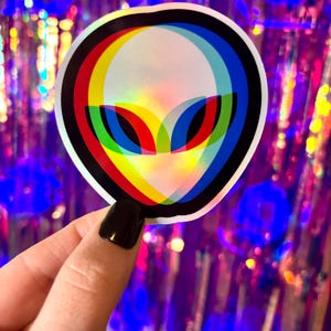 May include: A round sticker featuring a stylized alien head with a 3D effect. The design uses red, yellow, blue, and green colors, outlined in black. The sticker is held against a shiny, purple background.