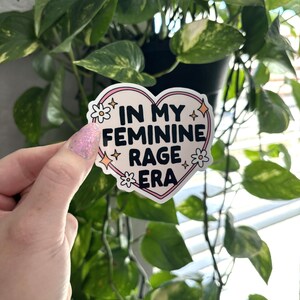 May include: A pink heart-shaped sticker with the text "In My Feminine Rage Era" in black lettering. The sticker has white flowers and glitter accents.
