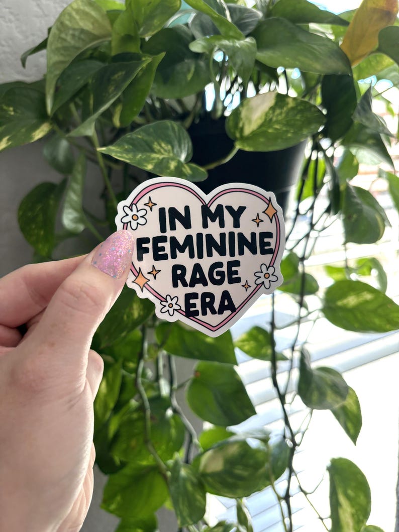 May include: A pink heart-shaped sticker with the text "In My Feminine Rage Era" in black lettering. The sticker has white flowers and glitter accents.