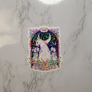 May include: A holographic sticker featuring a white cat with a crescent moon above it. The cat is surrounded by candles and flowers. The sticker has the text "XIII THE PROTECTOR" at the bottom.