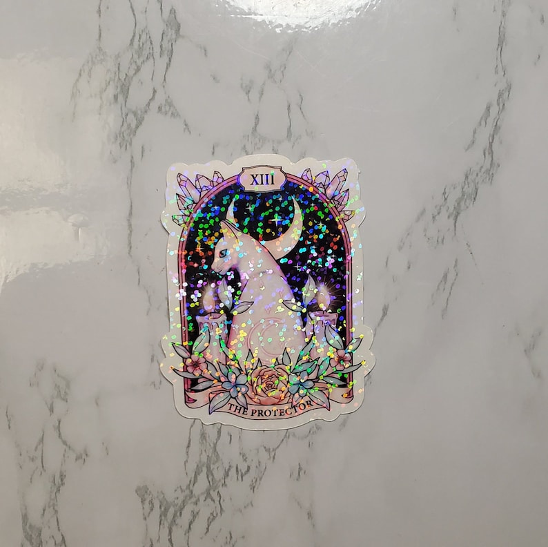 May include: A holographic sticker featuring a white cat with a crescent moon above it. The cat is surrounded by candles and flowers. The sticker has the text "XIII THE PROTECTOR" at the bottom.