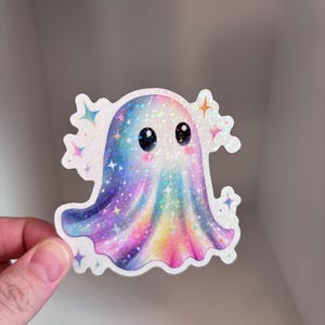May include: A holographic sticker of a cute ghost with a rainbow gradient design. The ghost has large, black eyes and is surrounded by sparkling star accents. The sticker has a white border and is held by a hand.