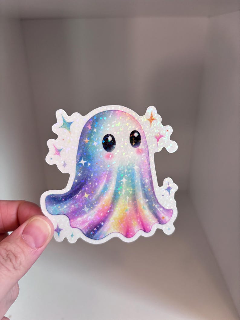 May include: A holographic sticker of a cute ghost with a rainbow gradient design. The ghost has large, black eyes and is surrounded by sparkling star accents. The sticker has a white border and is held by a hand.