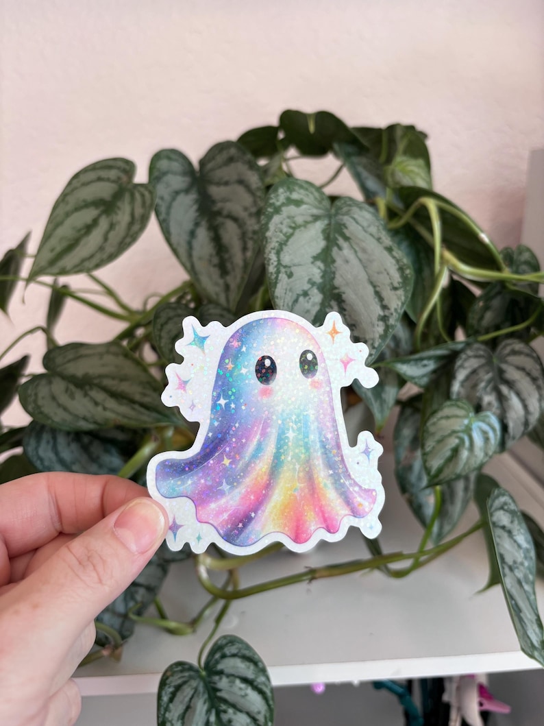 May include: A colorful sticker of a cartoon ghost with a rainbow gradient design. The ghost has a cute expression with large eyes and is surrounded by sparkling stars. The sticker is held in front of a green leafy plant.