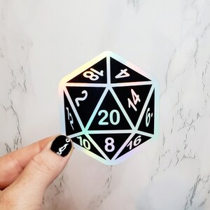 May include: A black and holographic D20 sticker with white numbers. The twenty-sided die is held by a hand with black nail polish. The sticker has a shiny, iridescent finish and is designed for tabletop gaming.