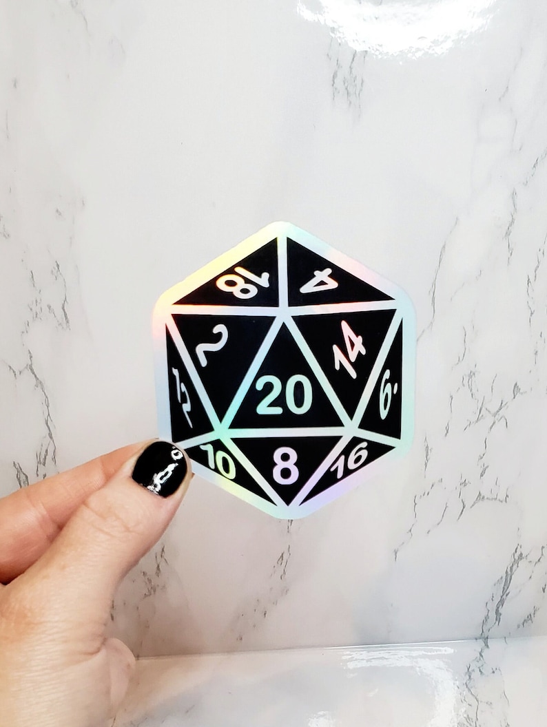 May include: A black and holographic D20 sticker with white numbers. The twenty-sided die is held by a hand with black nail polish. The sticker has a shiny, iridescent finish and is designed for tabletop gaming.