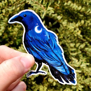 May include: A blue crow sticker with a crescent moon and stars on its feathers. The sticker is on a green leafy background.