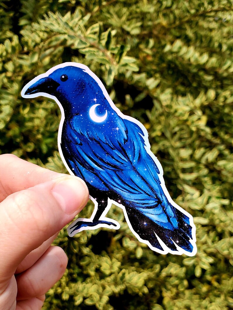 May include: A blue crow sticker with a crescent moon and stars on its feathers. The sticker is on a green leafy background.